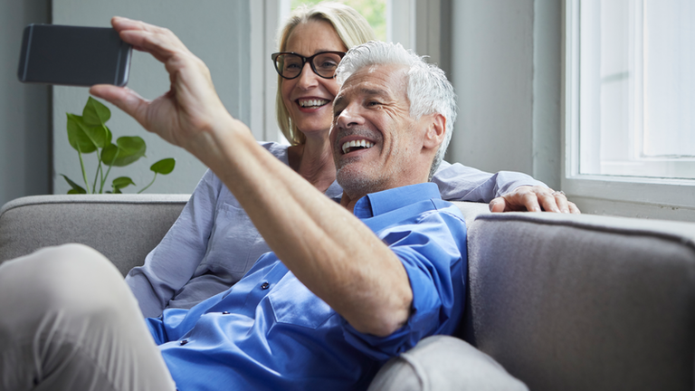 Happy mature couple sitting on couch at home taking a selfie